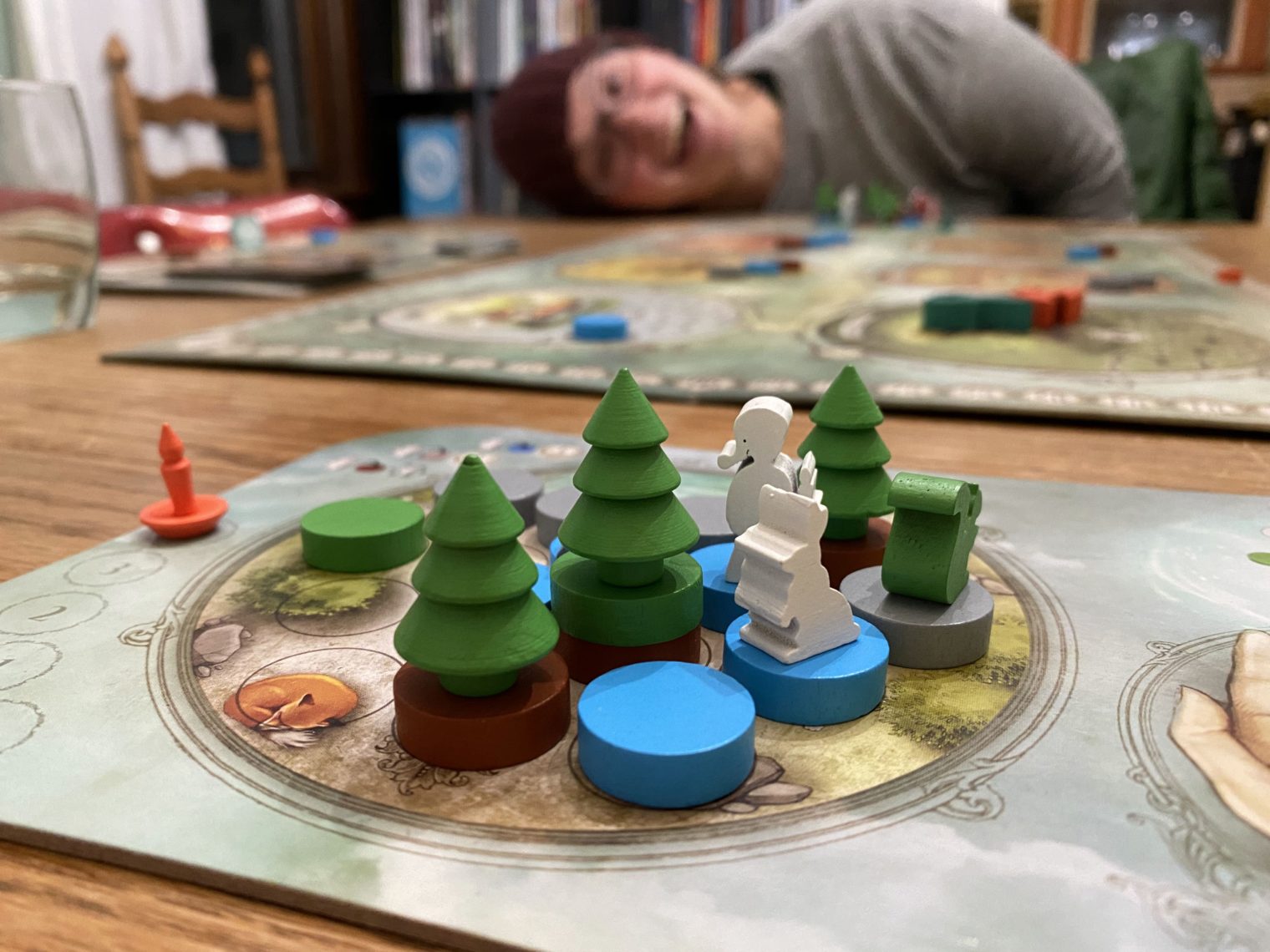 Boardgame set up in foreground. Person leaning over smiling in background slightly out of focus.
