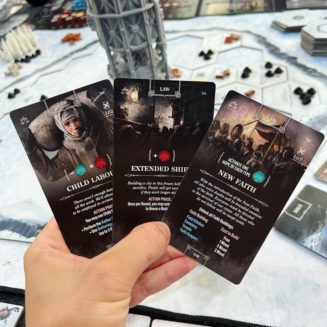 A hand holding three cards from Frostpunk: The Board Game, with a table view of the board game in the background.